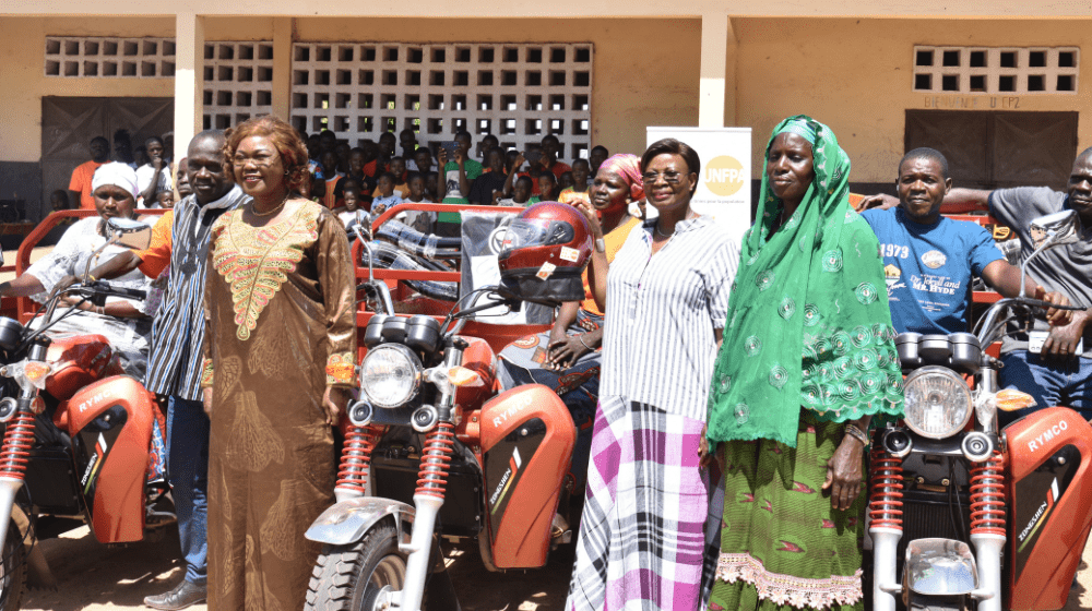 L'image montre un groupe de personnes debout devant plusieurs motocyclettes à trois roues (tricycles motorisés), alignées. Les motos sont de couleur rouge vif avec des détails noirs et portent le logo "RYMCO" sur le côté. Ces véhicules sont remis lors d'une cérémonie officielle