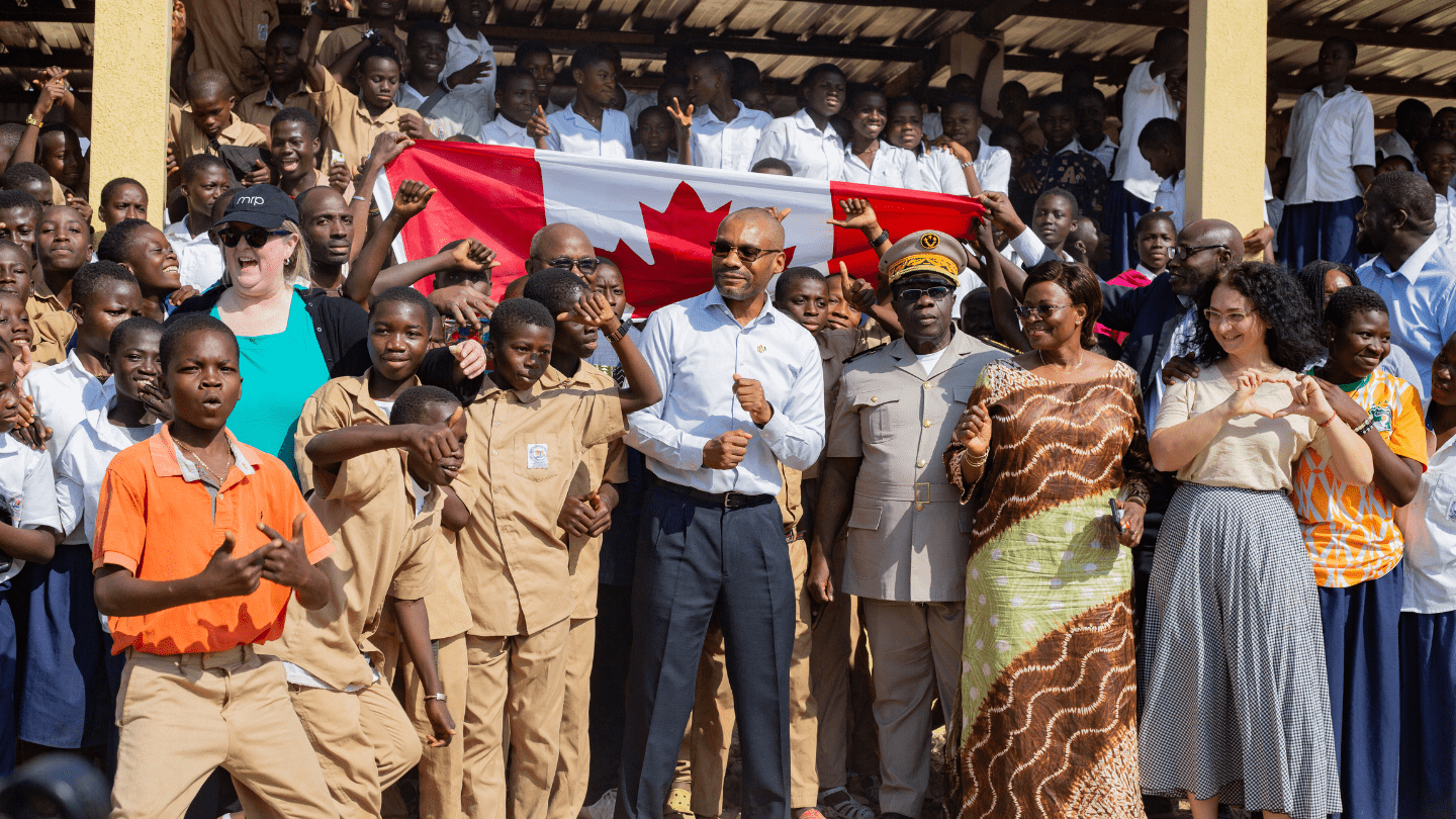 Une grande foule d’élèves en uniforme scolaire, accompagnés d’adultes, est rassemblée en extérieur sous une structure couverte. Plusieurs élèves portent des tenues kaki pour les jeunes garçons et blanche pour les filles.  Au centre, un homme vêtu d’une chemise blanche et de lunettes de soleil, entouré de plusieurs personnalités. À sa droite, un officier en uniforme beige et une femme en pagne traditionnel ivoirien participent également En arrière-plan, un drapeau canadien est tenu par des élèves