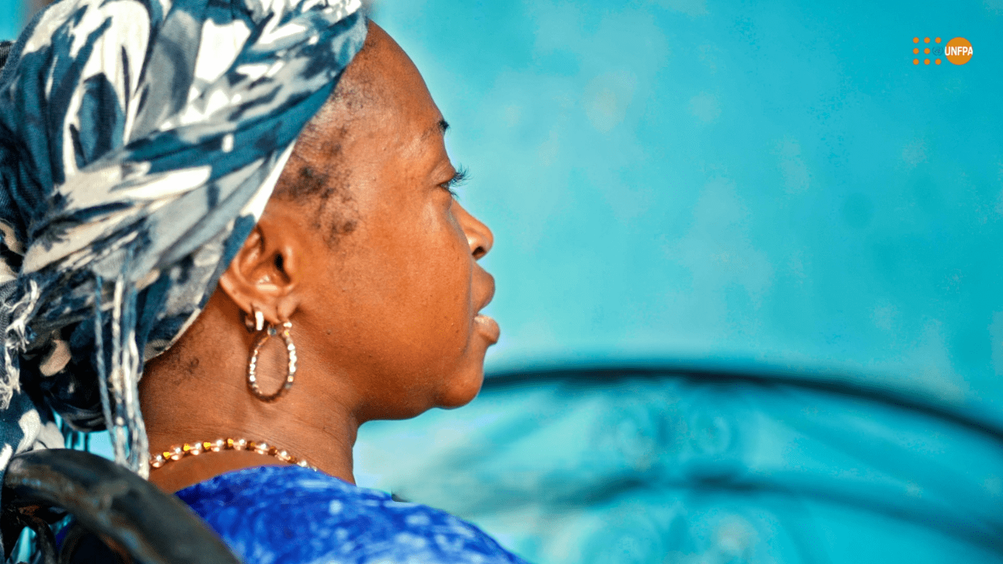 Portrait en profil d'une femme portant un foulard bleu et blanc, assise sur un lit en fer forgé, avec un fond bleu uni. Il s'agit de Kadi Koné (nom d’emprunt), une femme guérie de la fistule obstétricale après 10 ans de souffrance.