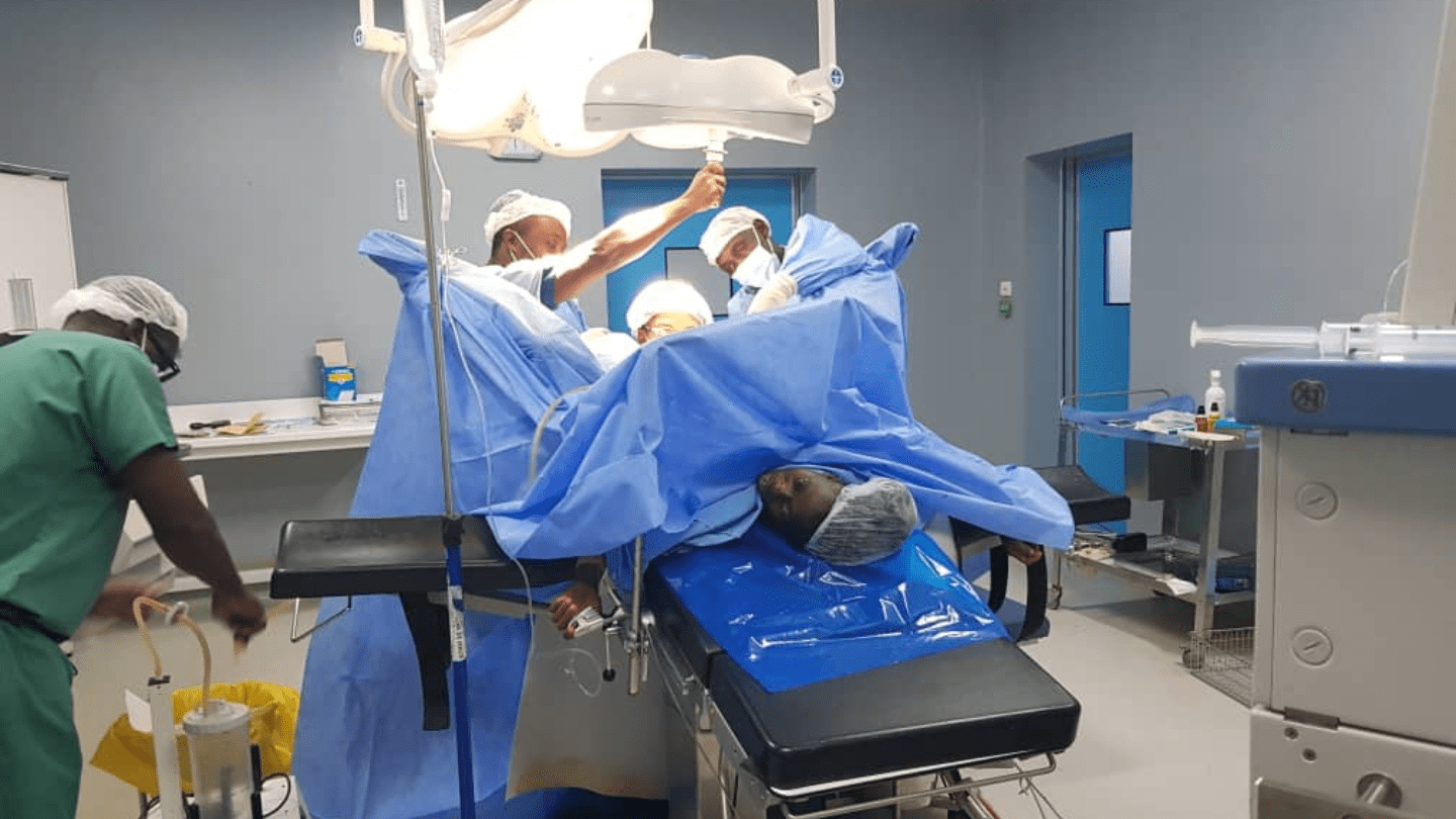 Une équipe chirurgicale composée de plusieurs médecins et infirmiers procède à une intervention de réparation de fistule obstétricale dans une salle d’opération stérile. Une patiente est allongée sur la table d’opération, entourée de champs opératoires bleus et de matériel médical